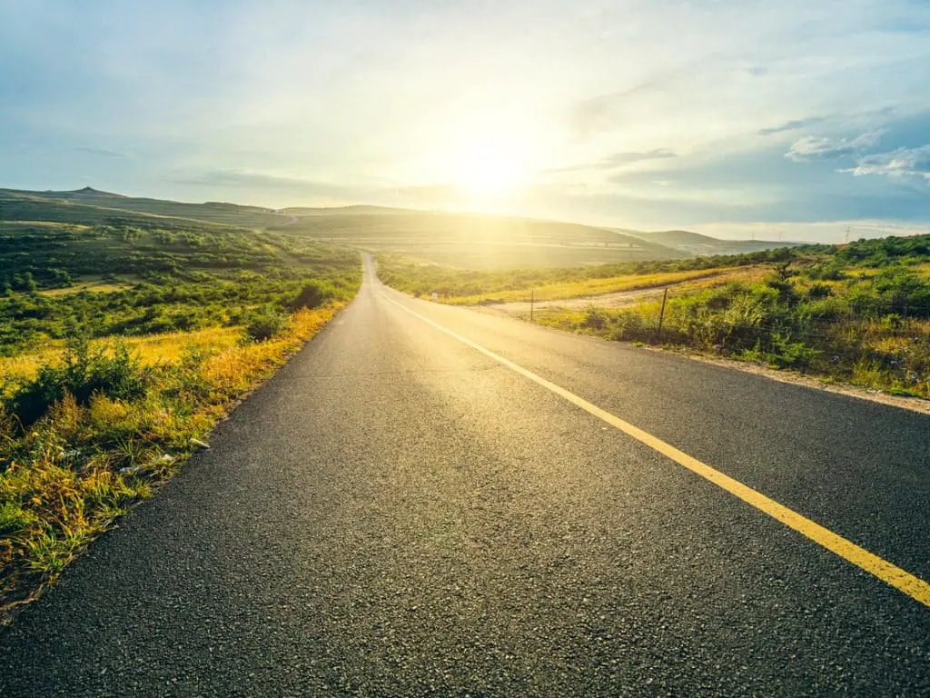 Photo of long two-lane road leading to sun rising over green hills