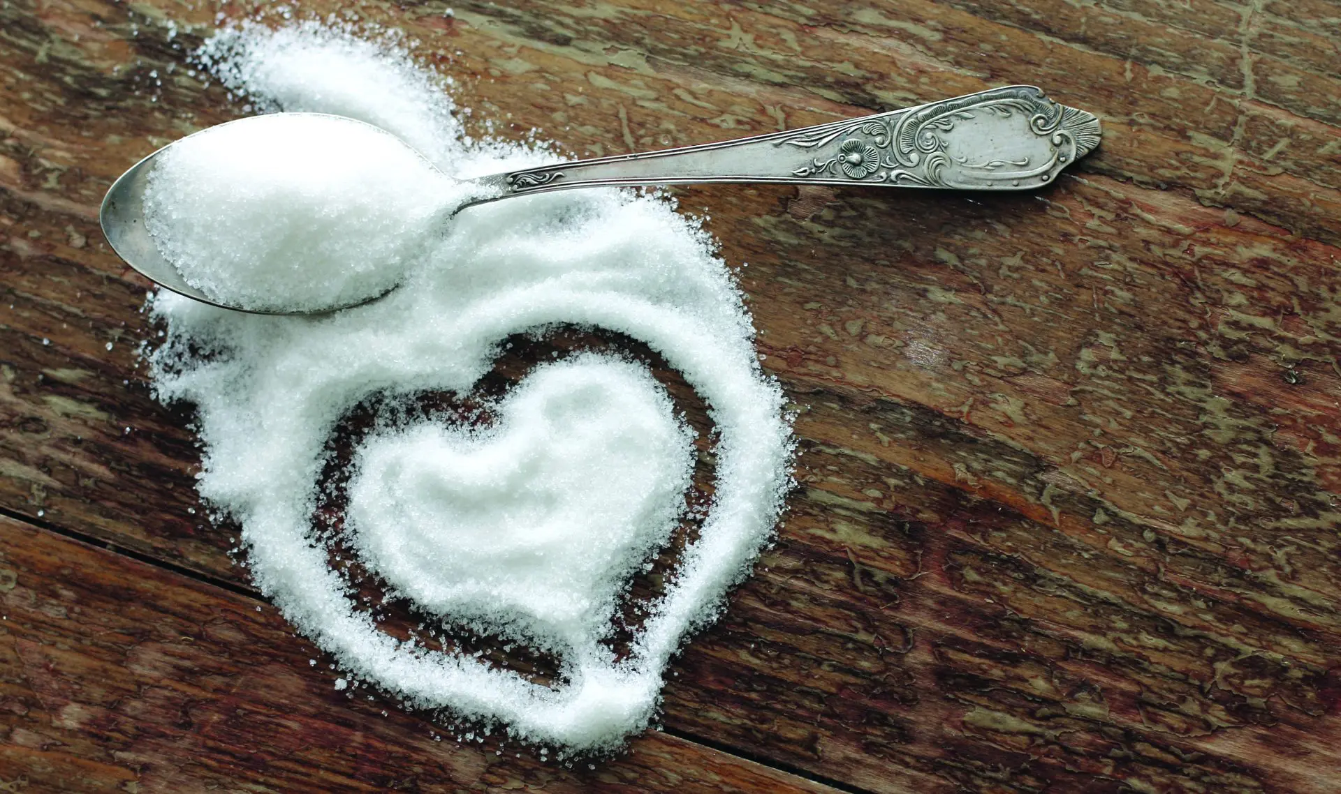Spoon full of sugar with a heart drawn in spilled sugar on a wooden table